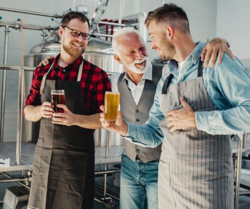 Three men holding glasses of beer, standing in a brewery.